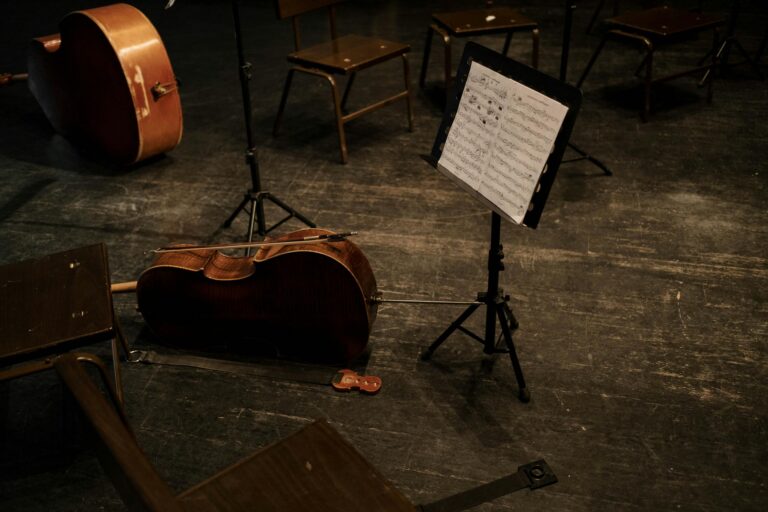 A cello rests on an empty stage with a music stand and scattered chairs, evoking classical music ambiance.